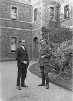 Image of two men, one in soldiers uniform, standing in front of bluestone building