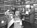 Photograph showing three women and a man at work in a library room lined with shelves of books. Several baskets of books are stacked in the centre of the room. 